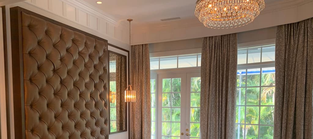 A bedroom wall of floor-to-ceiling tufted leather under a crystal chandelier, beside full-height drapes and French doors open to palms.