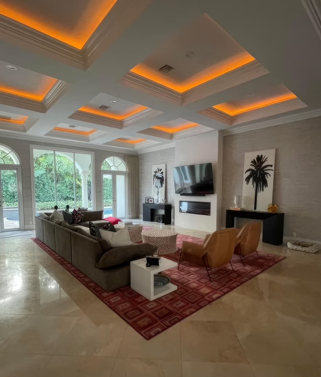 A great room with a coffered ceiling washed in amber cove light, arched windows and a sectional on a red patterned rug.